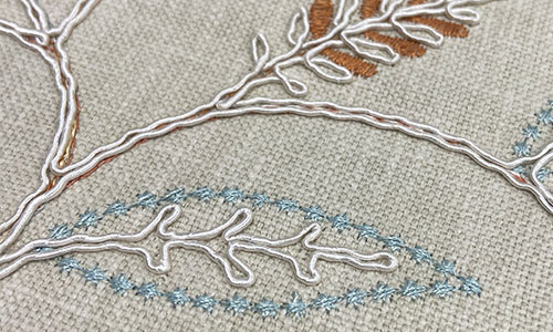 Detail of delicate embroidered leaves in light blue, white and deep orange on a visually textured beige background.