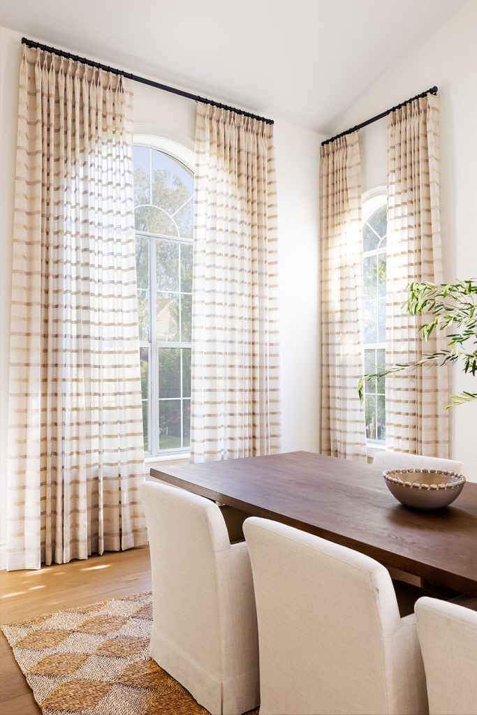 A dining room featuring two different sizs of arched windows with Monarch Pleat Sheer Drapery in a soft neutral wide stripe pattern.