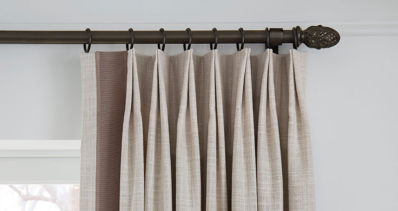 Decorative drapery hardware with a dark finish on light beige draperies with vertical trimming on the leading edge.