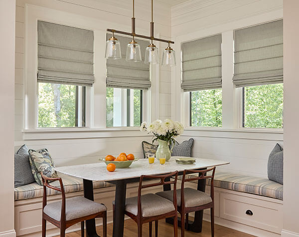 A kitchen dining nook featuring four Front Fold Romans Shades on thw windows over padded bench seating.