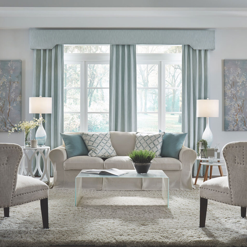 Pale blue Belvedere Cornice top treatment and Pleated Drapery over a wall of windows in a light neutral living room.