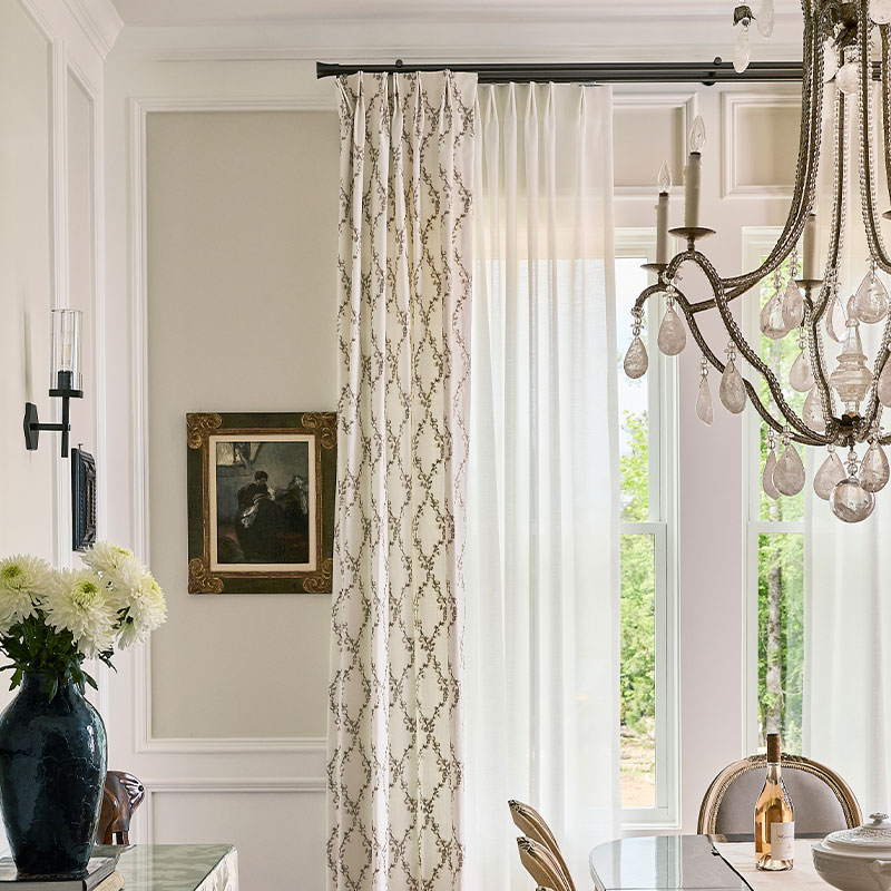 An elegant dining room featuring a whindow with white sheers and embroidered drapery on double traversing rods from Paris Texas Hardware.
