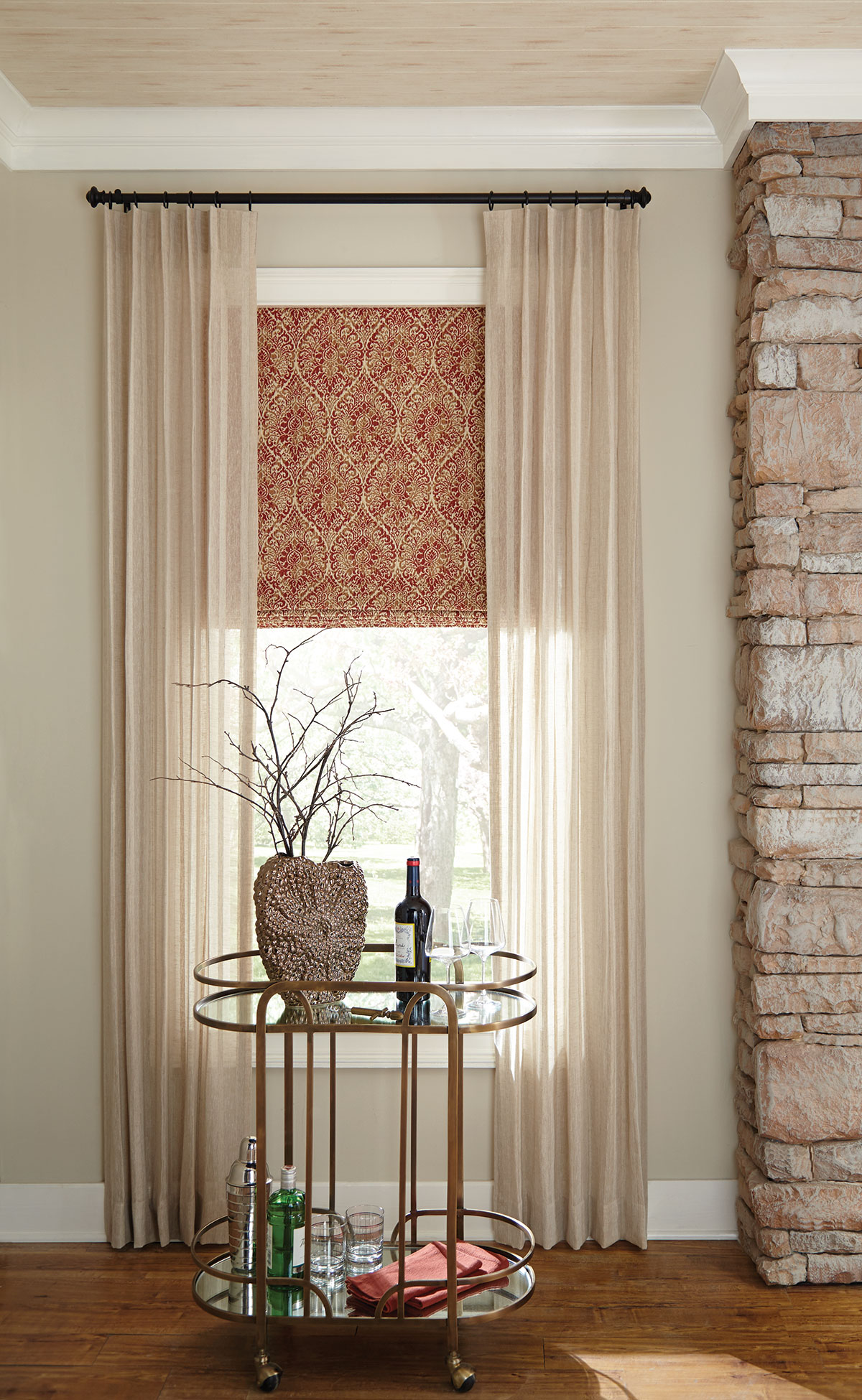 Sheer neutral draperies on a window over a flat Roman shade with a delicate metallic gold pattern on red fabric.