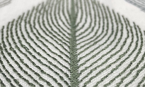 Detail of lines of green embroidery in the shape of a leaf on an off-white background.